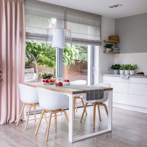 Gray roman shades and a pink curtain on big, glass windows in a modern kitchen and dining room interior with a wooden table and white chairs