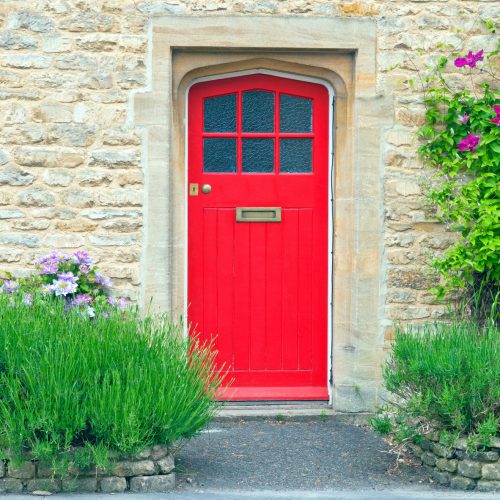 Bright red wooden doors in an old traditional English stone house, surrounded by climbing pink clematis on the golden wall, lavender in the front garden .